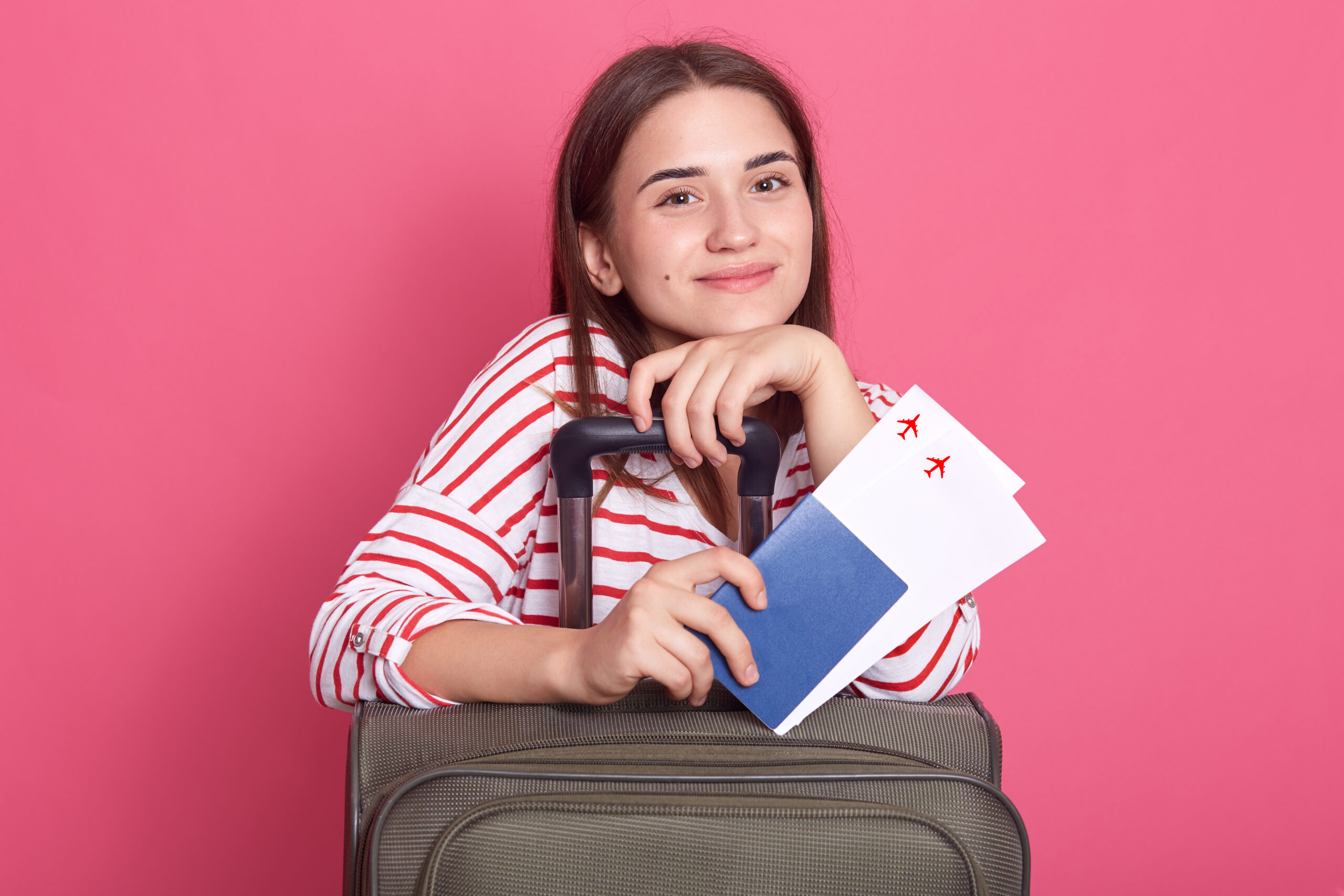Jeune femme souriante, appuyée sur une valise, tenant deux billets d'avion et un passeport, sur fond rose.