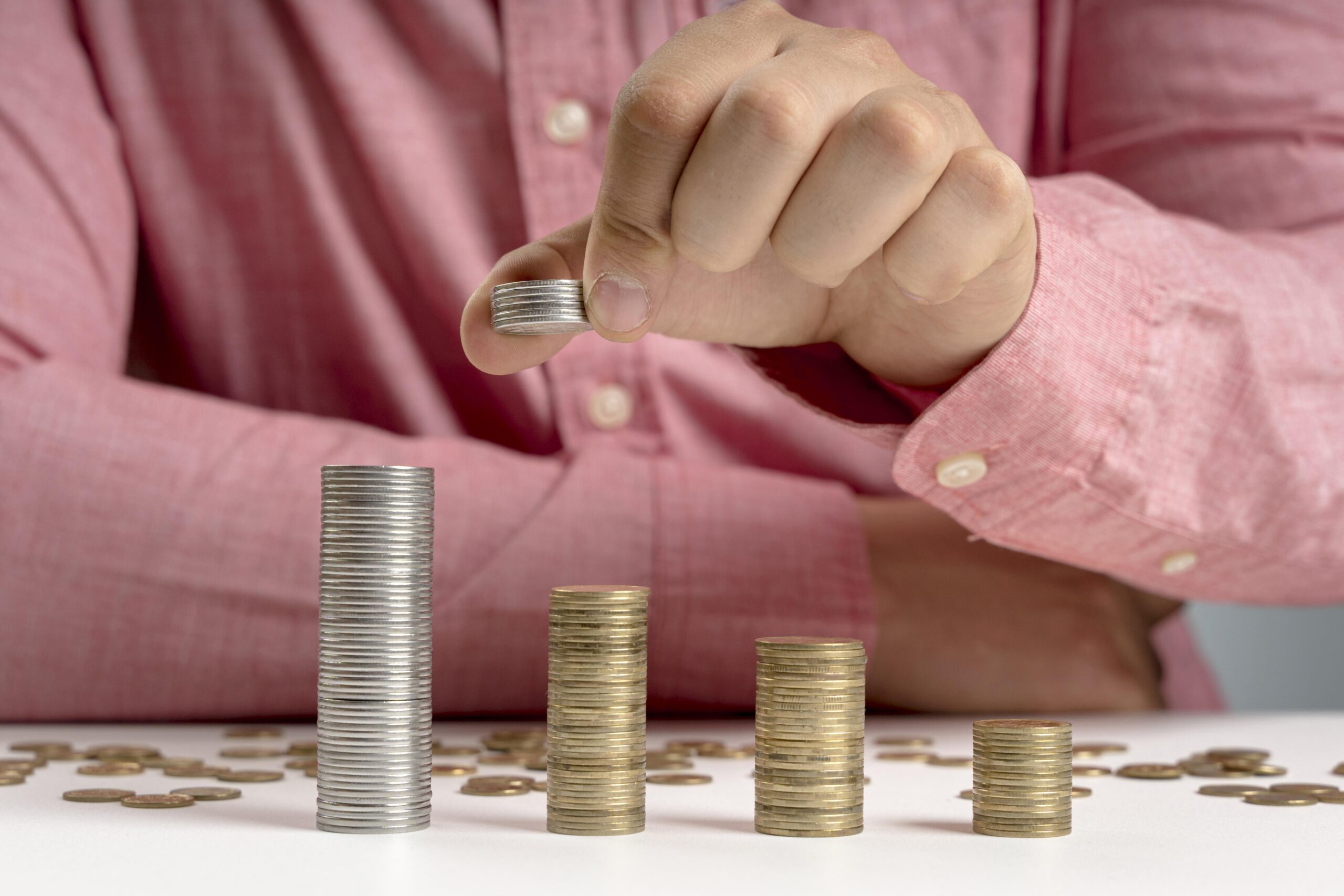Une personne vêtue d'une chemise rose empile des pièces de monnaie sur une surface blanche, créant ainsi quatre piles ascendantes de pièces, la plus haute se trouvant à gauche et la plus courte à droite. La main de la personne ajoute des pièces à une pile.