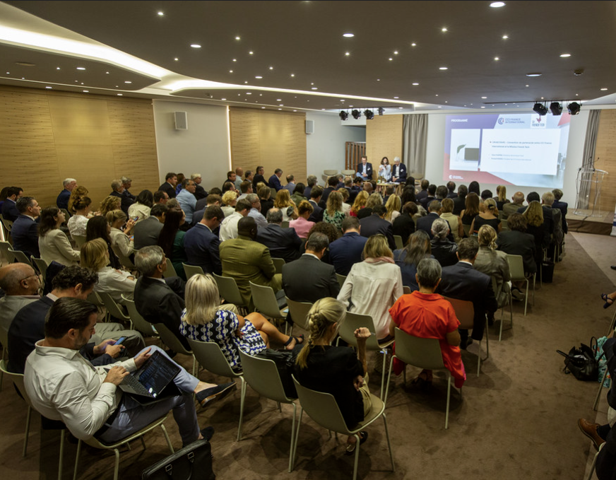Un grand groupe de personnes assises dans une salle de conférence écoute un panel d'orateurs à l'avant, avec une présentation projetée sur un écran derrière eux. Le public semble engagé et la salle est bien éclairée.